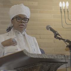 Rabbi Tamar Manasseh stands before a podium and microphone addressing a group of people out of frame. She is wearing all white attire at her ordination.
